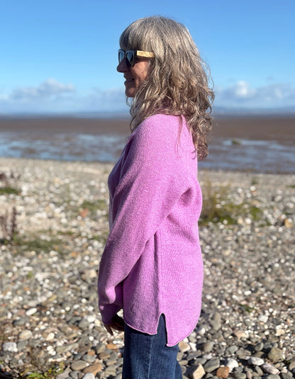 A woman with long gray hair and sunglasses stands in profile on a pebble beach, wearing a fuzzy lavender-pink knit cardigan and dark blue jeans, with water and a blue sky in the background.