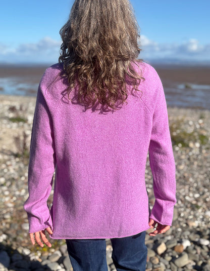 A person standing on a rocky beach, viewed from behind, wearing a long-sleeve knit sweater in a heathered light purple/lavender color and dark blue jeans.