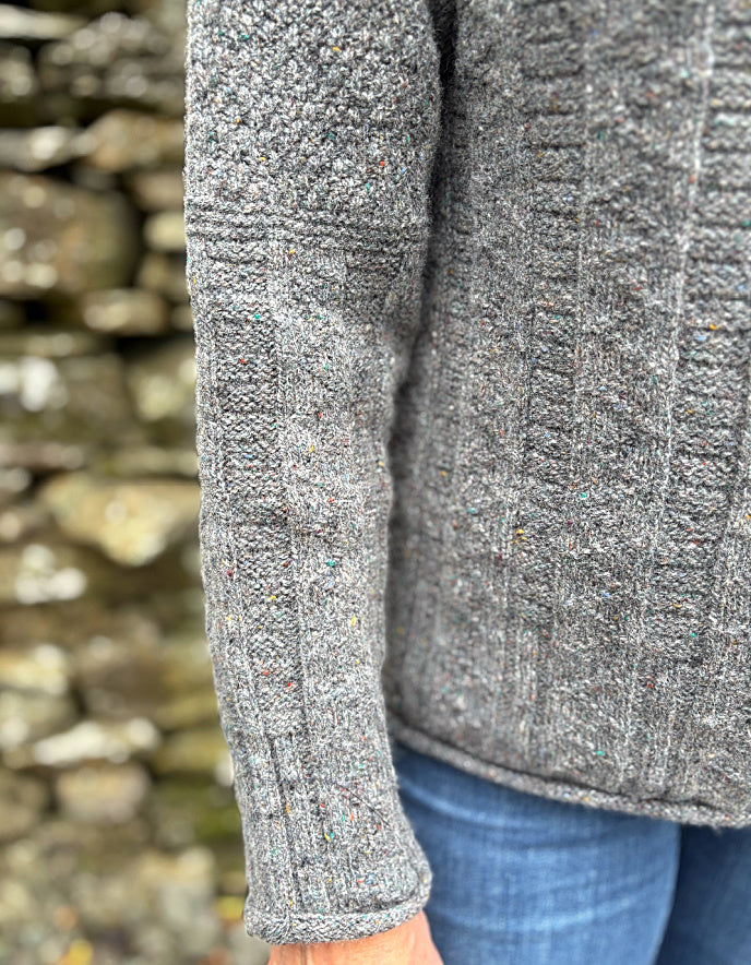 Close-up of a person wearing a gray textured sweater with a stone wall background