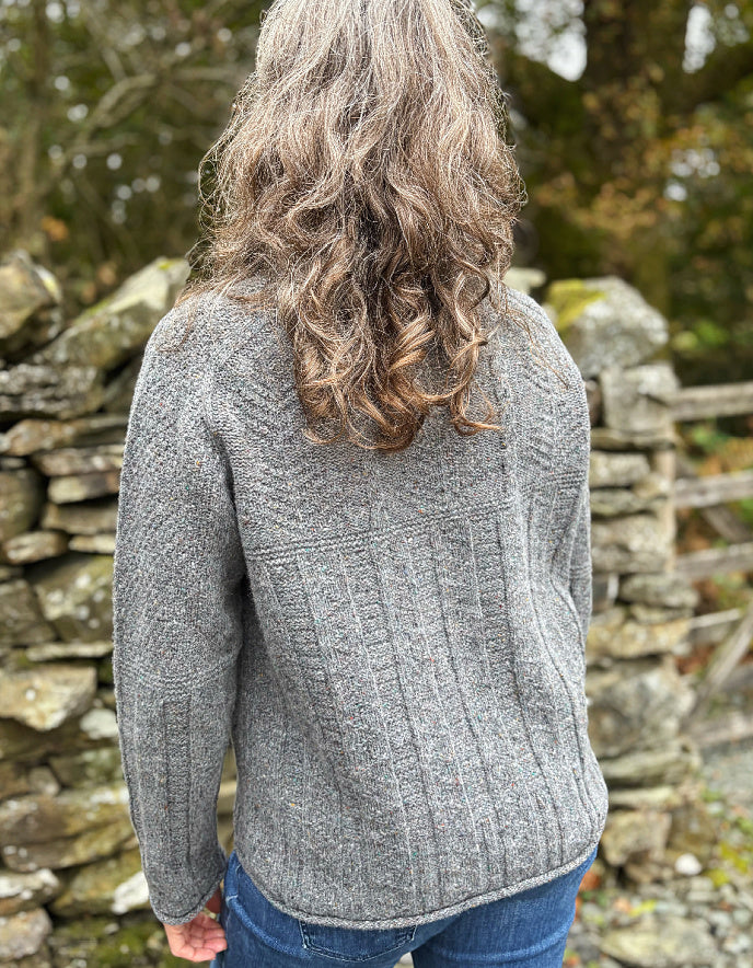 Person wearing a gray knitted sweater standing in front of a stone wall.