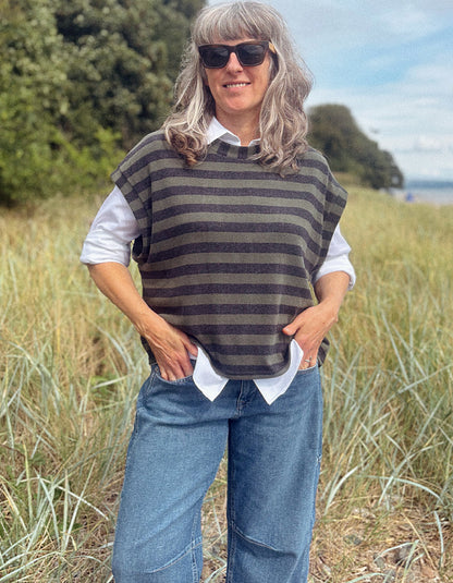 A woman wearing a dark gray and brown striped knit sweater vest over a white collared shirt and blue jeans, standing outdoors in a grassy field.