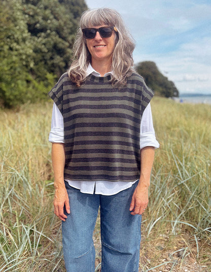 A woman with gray hair and sunglasses stands outdoors in a grassy field, wearing a dark gray and brown striped knit sweater vest over a white collared shirt and blue jeans.