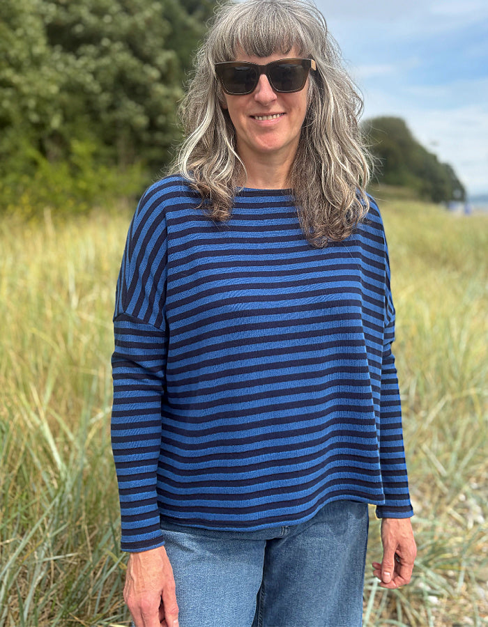 A woman with gray hair and sunglasses wearing a blue and navy striped long-sleeve knit top, standing outdoors in a grassy field.