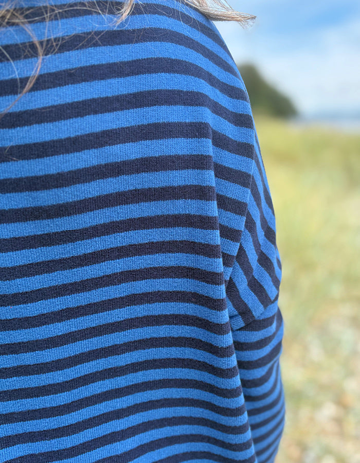 A close-up of a person wearing a blue and dark navy striped knit top, photographed outdoors with a blurred grassy field and sky in the background.