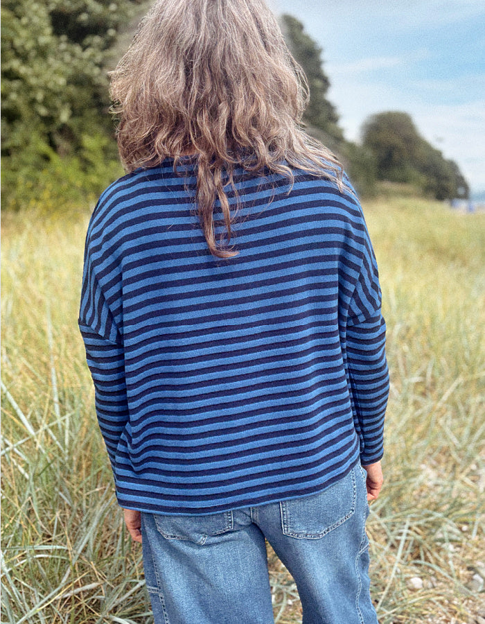 A child viewed from behind wearing a navy blue and dark navy striped long-sleeve shirt and blue jeans, standing in a grassy outdoor field.