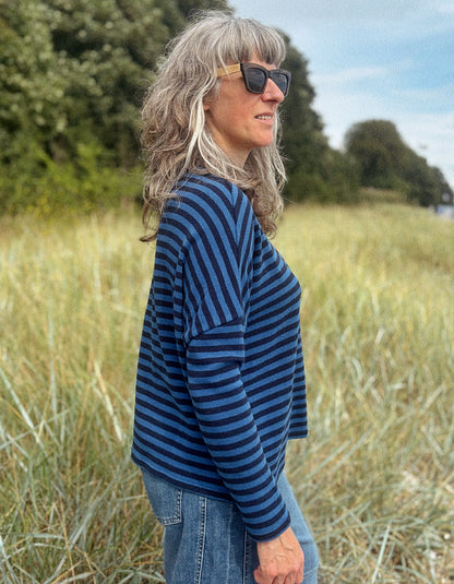 A woman with gray hair and sunglasses stands in a grassy field, wearing a navy and blue striped long-sleeve top and blue jeans.