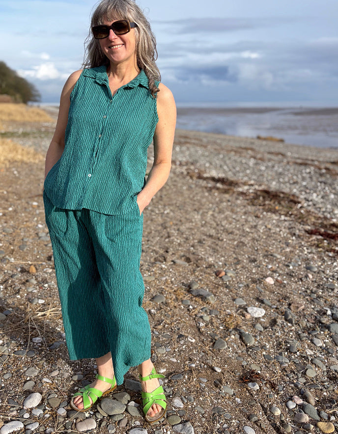 Woman in teal outfit standing on a pebbly beach with ocean and sky in the background