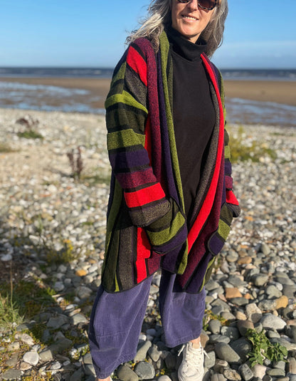 A woman standing on a rocky beach wearing a long striped cardigan with red, green, purple, and brown bands over a black turtleneck and loose purple pants.