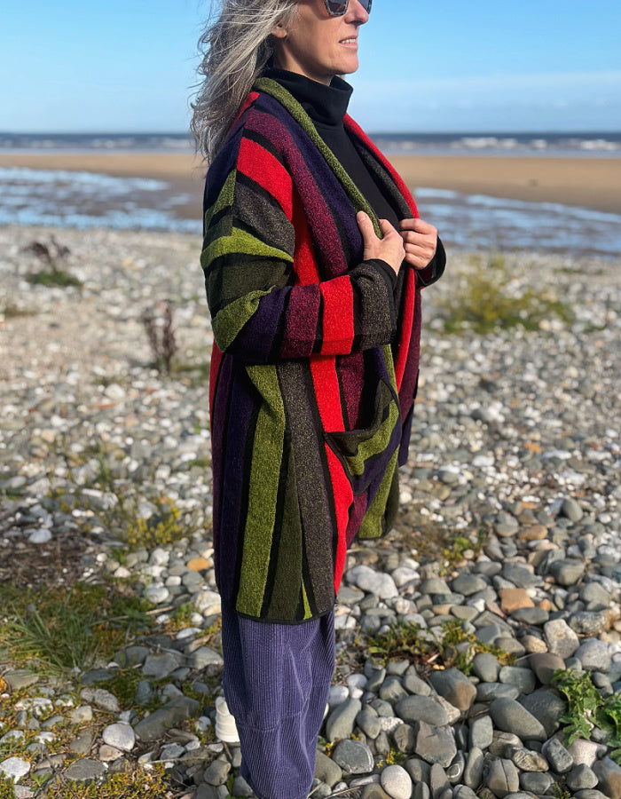 A woman standing on a rocky beach wearing a long striped cardigan with bold vertical stripes in olive green, red, purple, and black over a black turtleneck and purple pants.