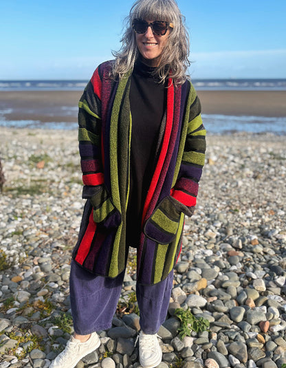 A woman standing on a pebble beach wearing a long striped cardigan with olive green, red, purple, and black stripes over a black turtleneck and loose purple sweatpants.