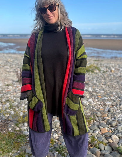 A woman standing on a pebble beach wearing a long open-front striped cardigan with bold vertical bands of olive green, red, purple, and black over a black turtleneck and loose gray pants.