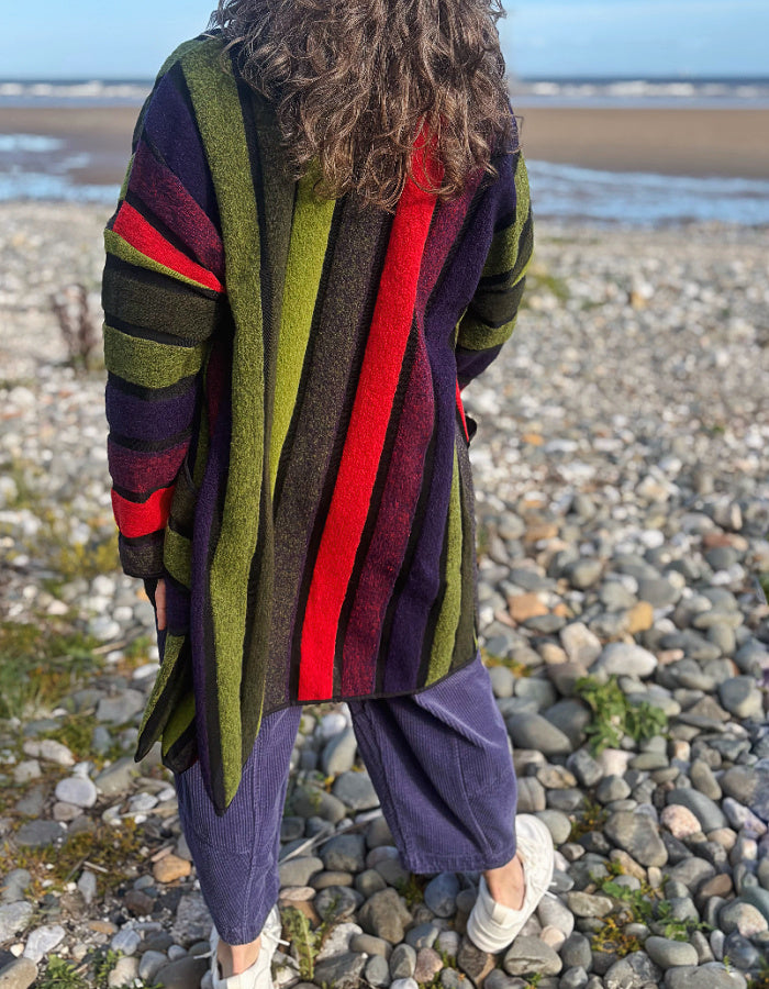 A person standing on a pebble beach wearing a long striped cardigan with vertical stripes in olive green, dark purple, red, and brown, paired with loose purple pants and white sneakers.