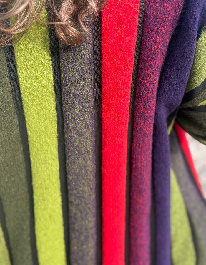 Close-up of a striped knit garment with vertical bands of olive green, charcoal gray, bright red, and deep purple, with a person's hair visible at the top.