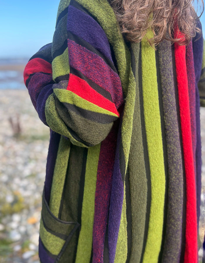 A close-up of a person wearing a thick, striped wool cardigan outdoors. The cardigan features vertical stripes of olive green, dark purple, red, and black, with horizontal stripes on the sleeve.