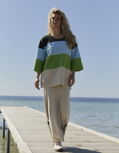 A woman walks on a wooden dock by the sea wearing a color-blocked knit top with horizontal stripes of black, light blue, white, green, and beige, paired with beige wide-leg pants.