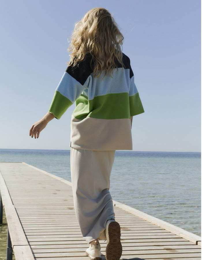 A woman walking away on a wooden dock by the sea, wearing a color-blocked short-sleeve top with black, light blue, green, and beige sections, paired with light beige wide-leg pants.