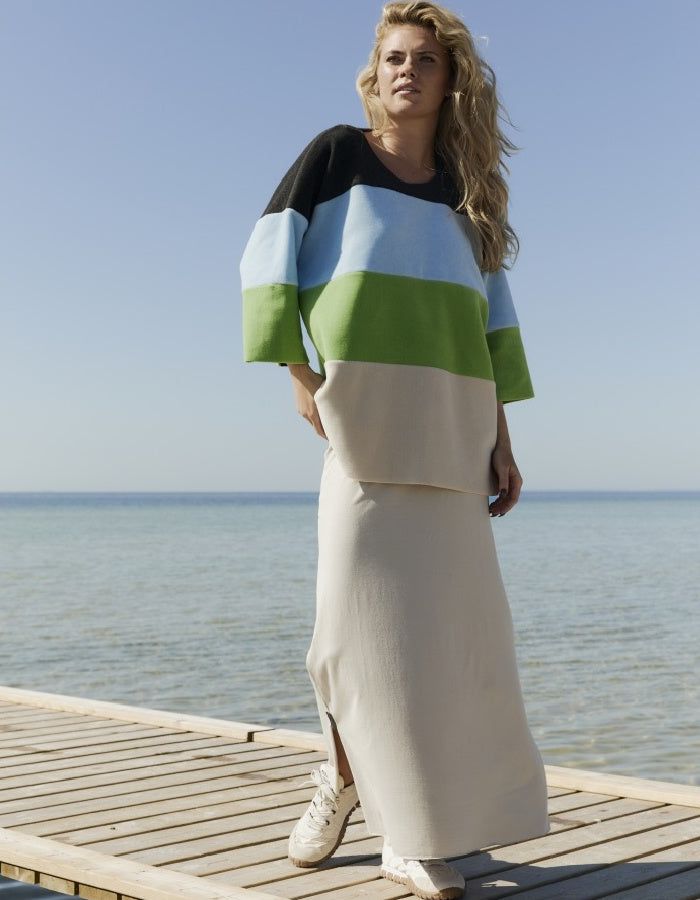 A woman stands on a wooden dock by the sea wearing a color-block knit top and a long cream skirt. The top has wide sleeves and horizontal color blocks of black, light blue, green, and cream.