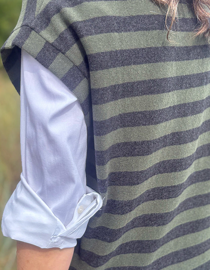 Close-up of a person wearing a sleeveless knit top with horizontal olive green and dark charcoal gray stripes, layered over a light blue button-up shirt with the cuff rolled up.