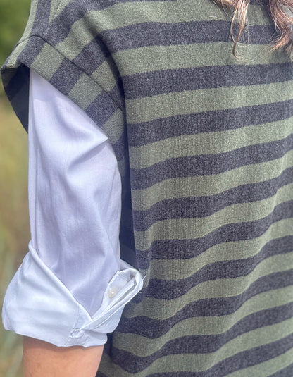 Close-up of a person wearing a sleeveless knit top with horizontal olive green and dark charcoal gray stripes, layered over a light blue button-up shirt with the cuff rolled up.