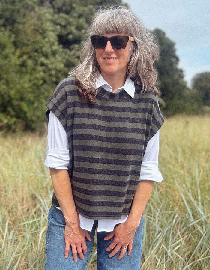 A woman with gray hair and sunglasses wearing a dark gray and olive green striped sleeveless sweater vest over a white collared shirt and blue jeans, standing in a grassy field.