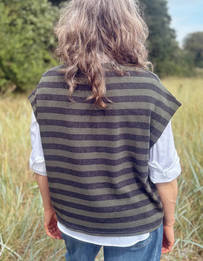 A person standing outdoors in a grassy field, viewed from behind, wearing a sleeveless knit top with horizontal dark olive green and charcoal gray stripes over a white shirt and blue jeans.