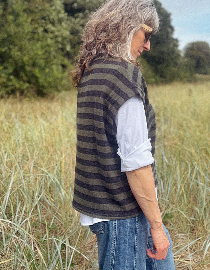 A woman standing in a grassy field, wearing a dark olive green and black striped knit sweater vest over a white long-sleeve shirt and blue jeans.