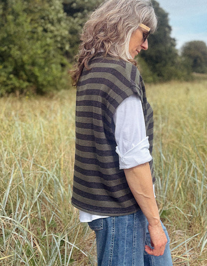 A woman standing in a grassy field, wearing a dark olive green and black striped knit sweater vest over a white long-sleeve shirt and blue jeans.