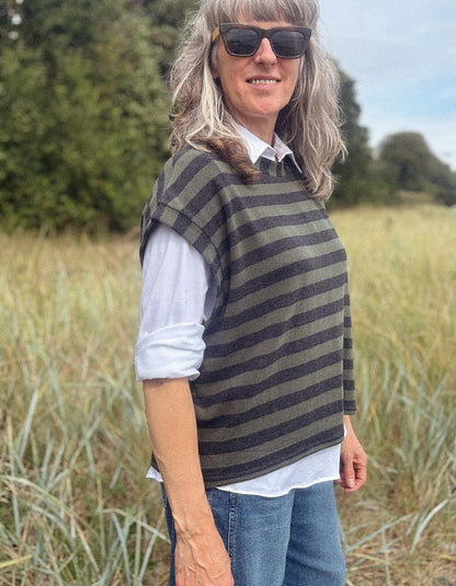 A woman wearing a dark olive green and black striped knit poncho over a white collared shirt and blue jeans, standing in a grassy field outdoors.
