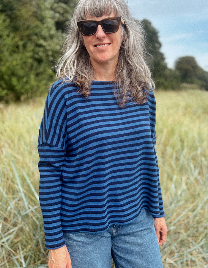A woman with gray hair and sunglasses wearing a navy blue and dark blue horizontally striped long-sleeve top, standing in a grassy outdoor field.