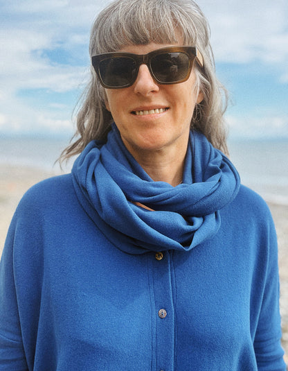 A woman with gray hair and sunglasses wearing a bright blue knit cardigan and matching blue infinity scarf, photographed outdoors against a soft-focus beach and sky background.