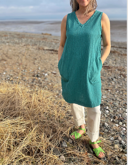 Woman in a teal sleeveless top and white pants standing on a pebbly beach.