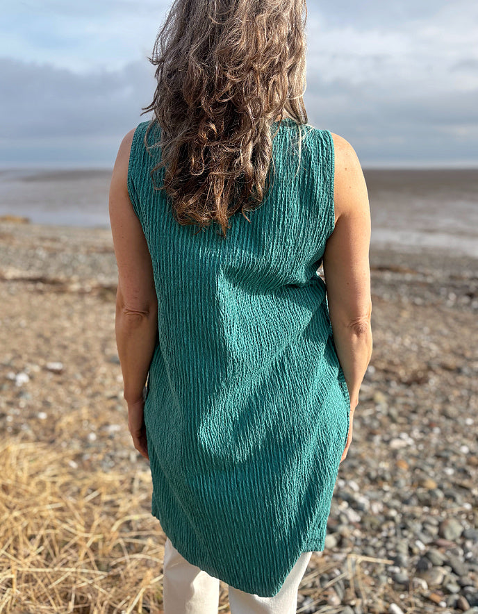 Person wearing a teal sleeveless dress standing on a pebbly beach with a cloudy sky.