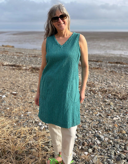 Woman in a teal sleeveless top and white pants standing on a pebbly beach.