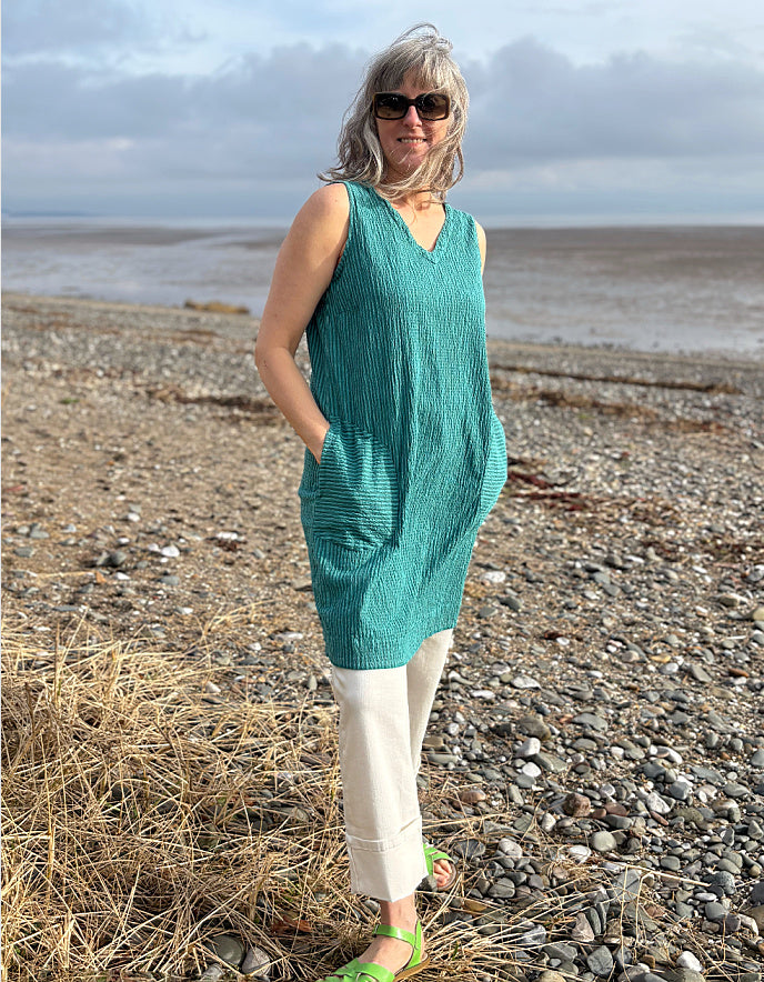Woman in a teal dress standing on a pebbly beach with water in the background
