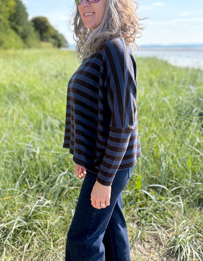 Woman wearing a striped sweater and sunglasses standing in a grassy field with a body of water in the background.