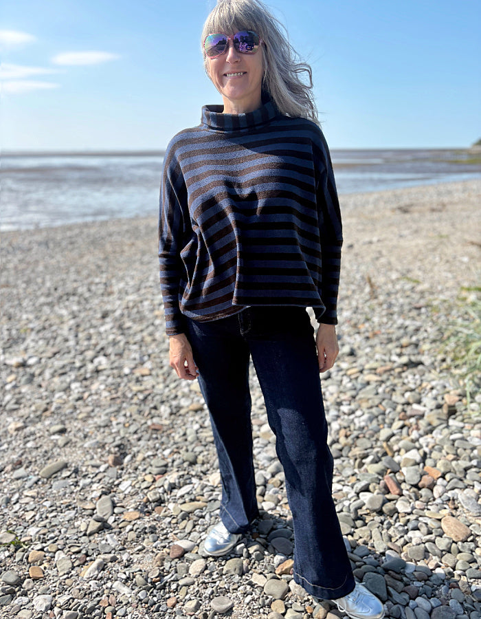 Person standing on a pebbly beach wearing a striped sweater and sunglasses.