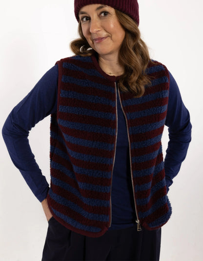 A woman wearing a navy blue long-sleeve shirt and a burgundy and navy striped fleece zip-up vest, paired with a burgundy knit beanie, against a plain white background.