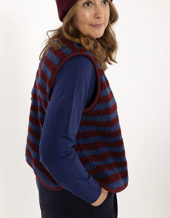 A woman wearing a striped knit vest over a navy blue long-sleeve top and a burgundy beanie, photographed against a plain white studio background.