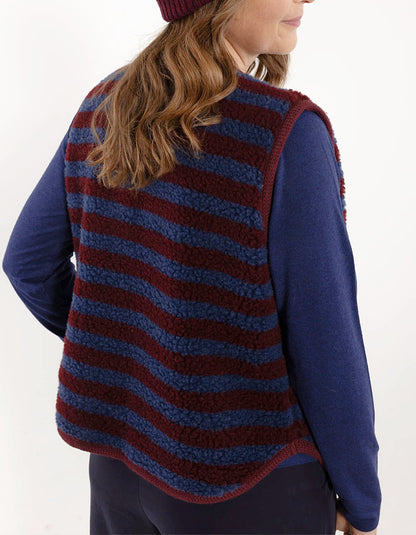 A woman shown from the back wearing a blue and burgundy striped sherpa fleece vest over a navy blue long-sleeve shirt, with a burgundy beanie partially visible.