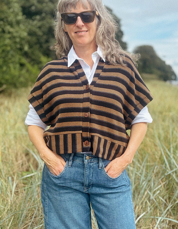 A woman wearing a brown and black striped knit poncho cardigan over a white collared shirt and blue jeans, standing in a grassy outdoor field.