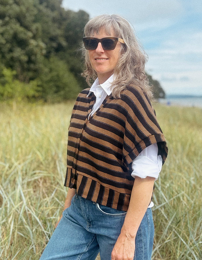 A woman with gray hair and sunglasses stands in a grassy outdoor field, wearing a brown and black striped knit poncho over a white collared shirt and blue jeans.