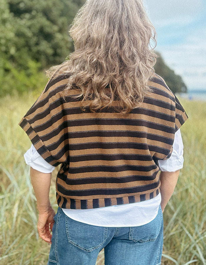 A person viewed from behind wearing a brown and black horizontally striped knit poncho-style top over a white shirt and blue jeans, standing outdoors in a grassy field.