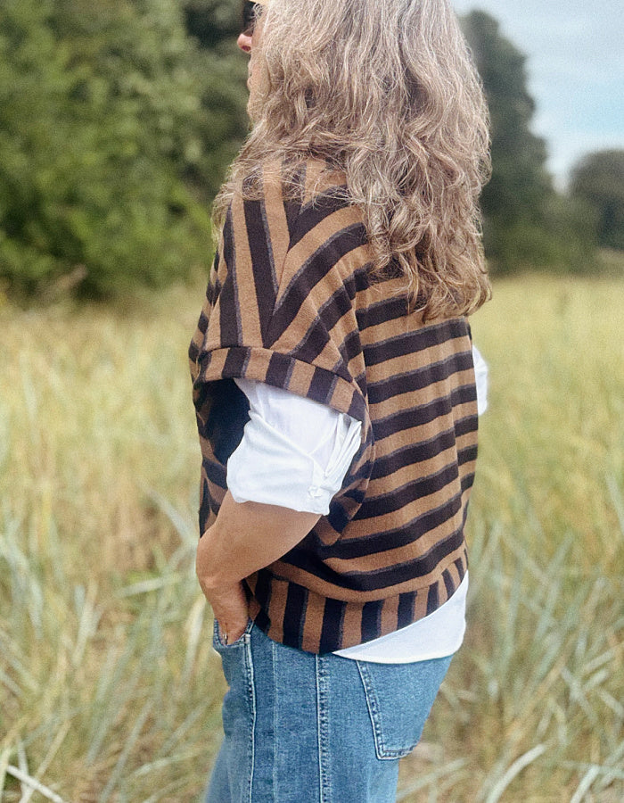 A woman standing outdoors in a grassy field, wearing a brown and black striped knit cardigan over a white top and blue jeans.