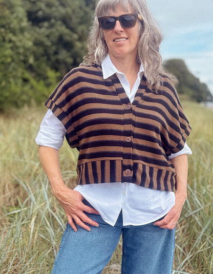 A woman wearing a brown and black striped knit poncho cardigan over a white collared shirt and blue jeans, standing outdoors in a grassy field.