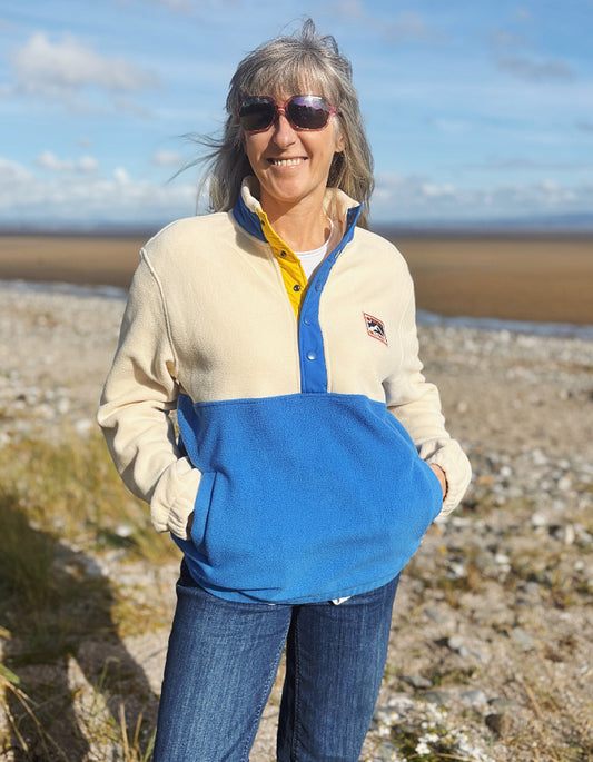 Woman wearing a two-tone fleece jacket with a blue front and beige back, standing on a beach.