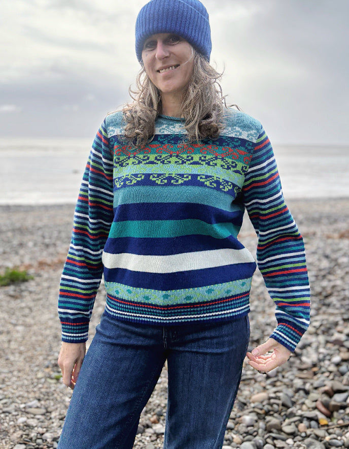 Person wearing a colorful patterned sweater and blue hat standing on a pebbly beach.