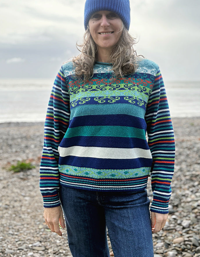 Person wearing a colorful striped sweater with a patterned design, standing on a pebbly beach.