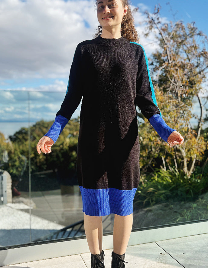 Woman wearing a black and blue colour block knitted sweater dress standing outdoors with trees and sky in the background