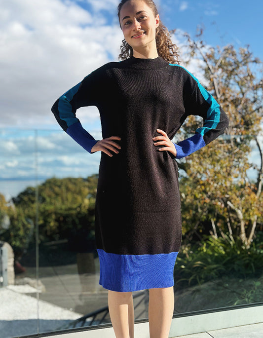 Woman wearing a black and blue colour block knitted sweater dress standing outdoors with trees and sky in the background