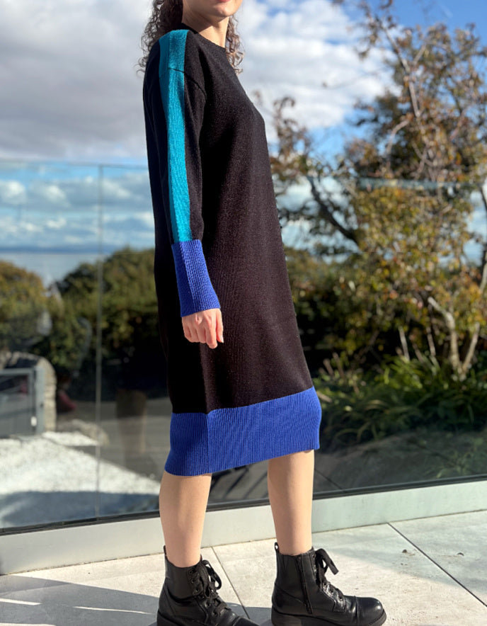 Woman wearing a black and blue colour block knitted sweater dress standing outdoors with trees and sky in the background